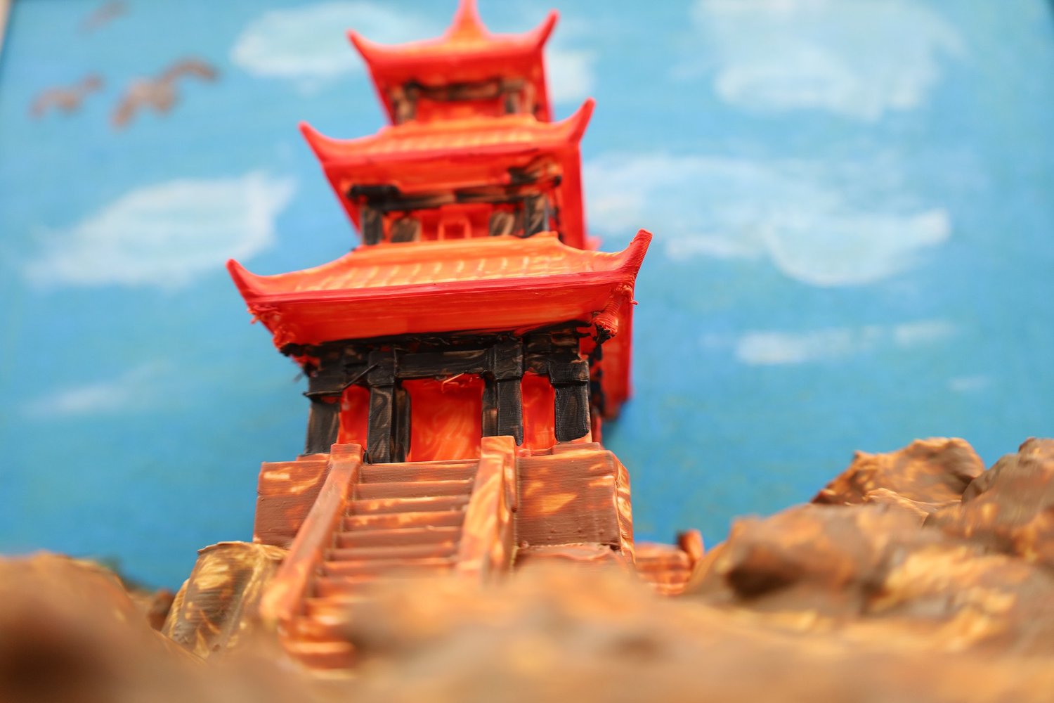 A bright red 3D-printed pagoda climbing a cliff against a painted blue sky